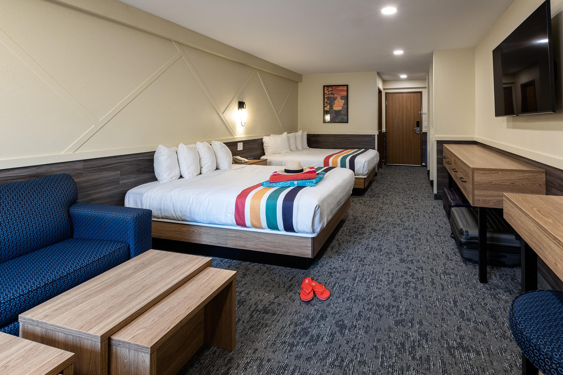 A hotel room with two double beds, a blue sofa, a wooden coffee table, a desk with a chair, and a flat-screen TV on the wall.