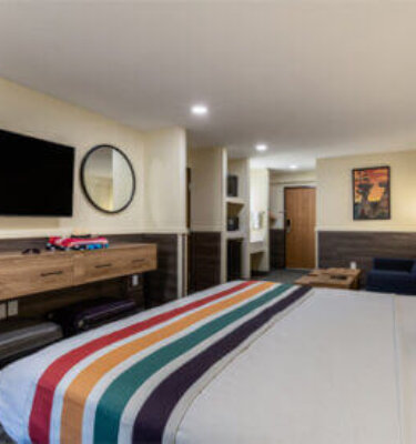 A wide-angle view of a hotel room with a bed, TV, desk, and mirror.