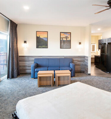 A wide view of a hotel room with a blue sofa, two wooden coffee tables, a bed, and a TV.