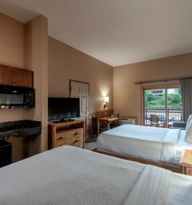 A hotel room with two double beds, a desk, a microwave, and a TV, with a balcony overlooking a green hillside.